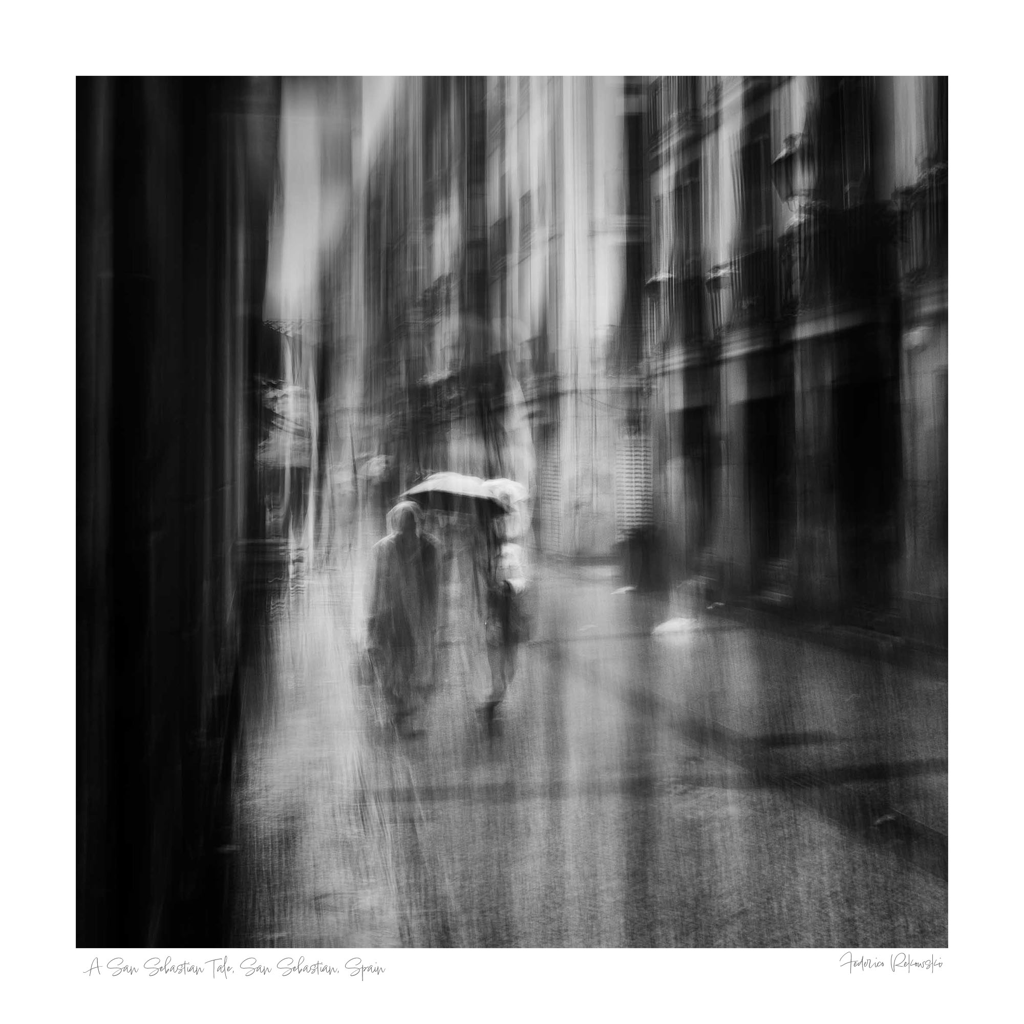 Black and white image of blurred figures under an umbrella on a rainy street in San Sebastian, Spain, creating an ethereal atmosphere.