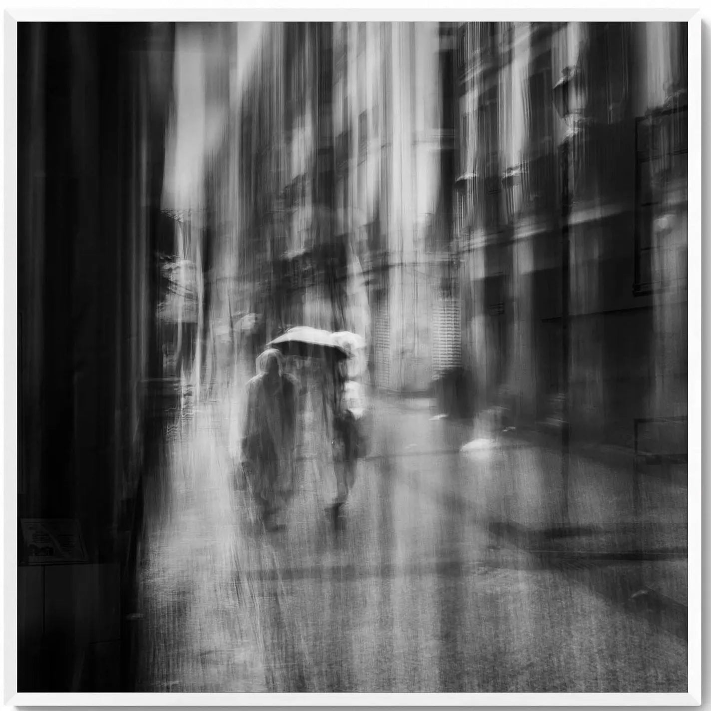 Black and white image of blurred figures under an umbrella on a rainy street in San Sebastian, Spain, creating an ethereal atmosphere.