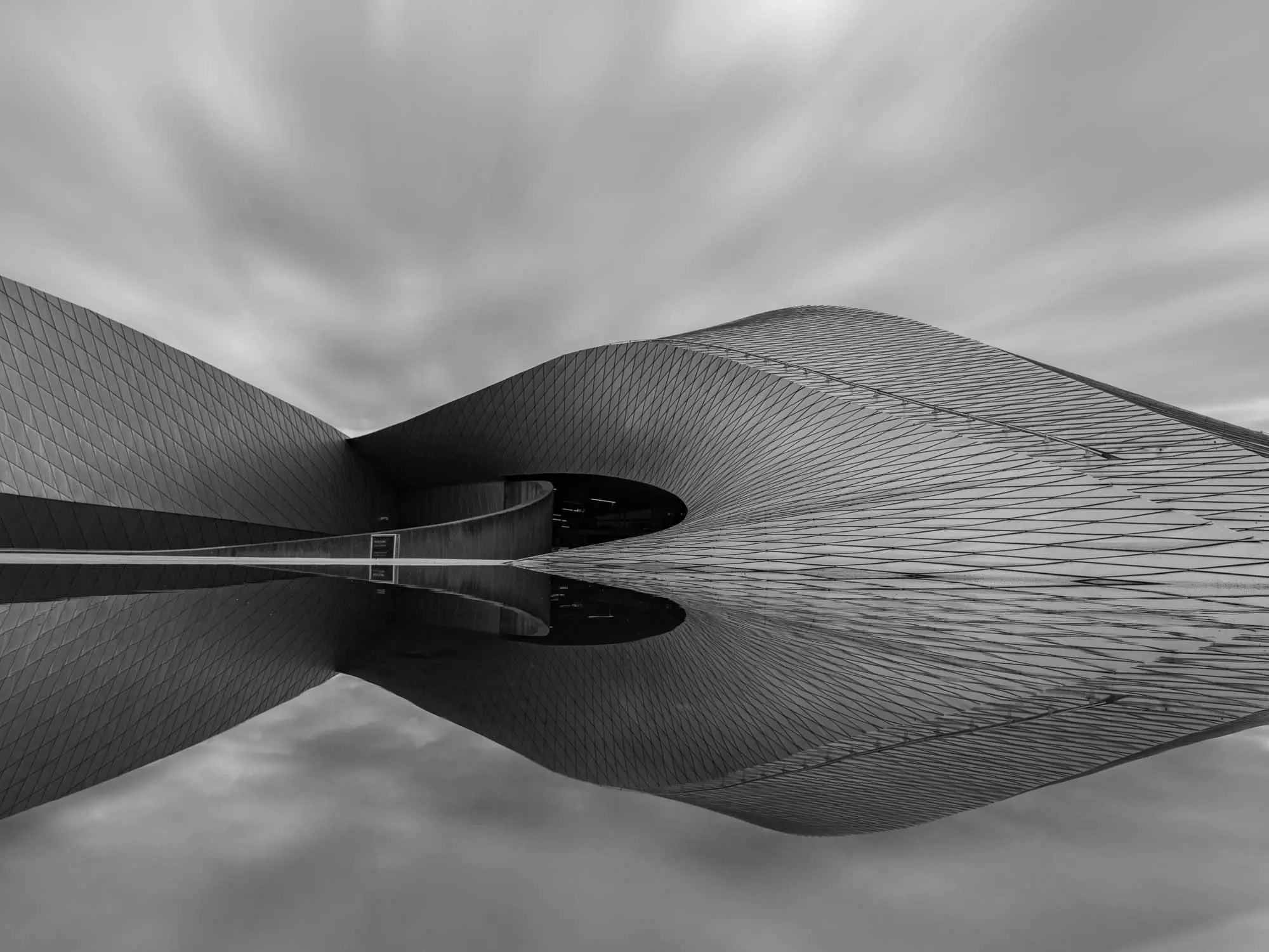 Black and white image of The Blue Planet Aquarium in Copenhagen with its reflective, wavy facade against a blurred sky.