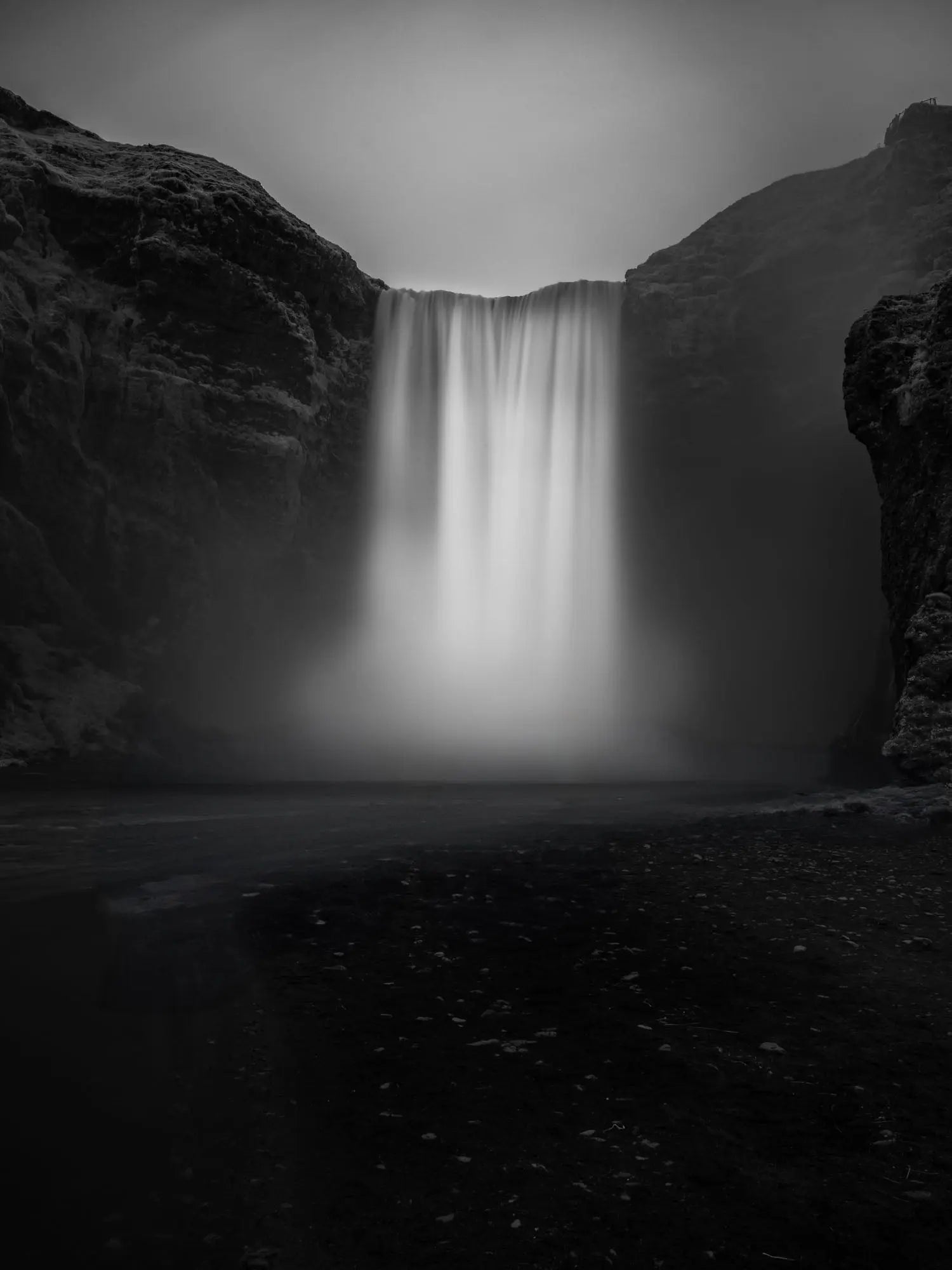 A long exposure photograph of Skógafoss waterfall in Iceland, rendered in stunning black and white, capturing the smooth flow of water between two dark cliffs.