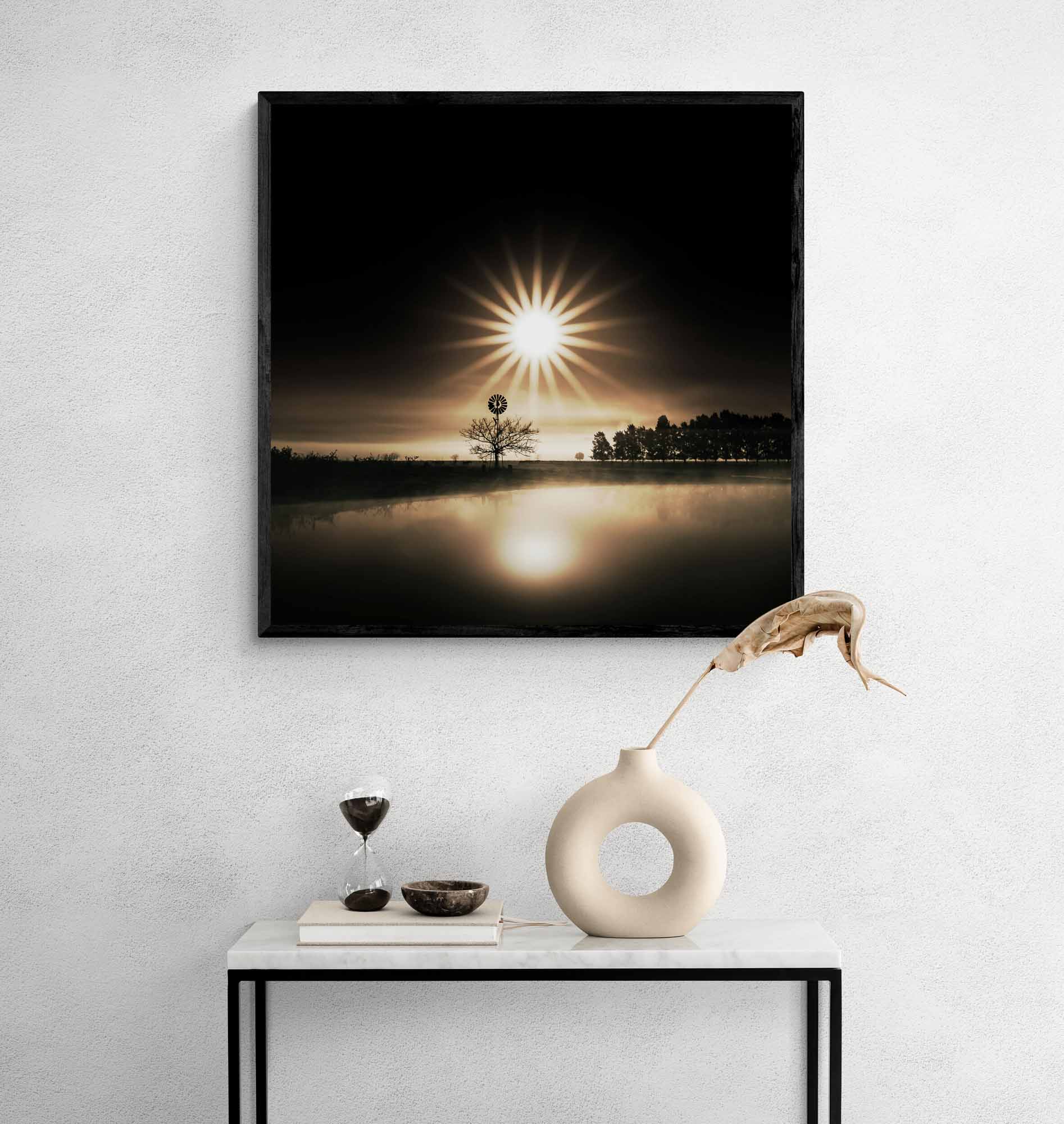 A serene sunrise with a lone tree silhouette against a bright sunburst, reflected on a calm water surface.