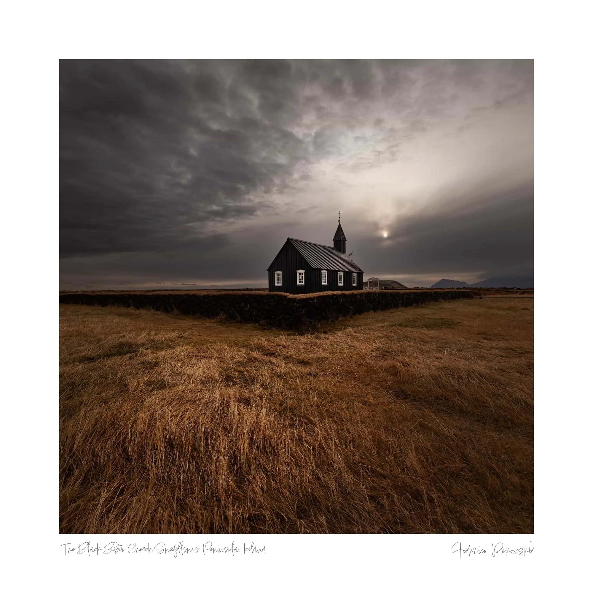 A dark, solitary church in the open Icelandic landscape with dry grass in the foreground and a dramatic, cloud-filled sky above, capturing a moment of quietude before a storm.
