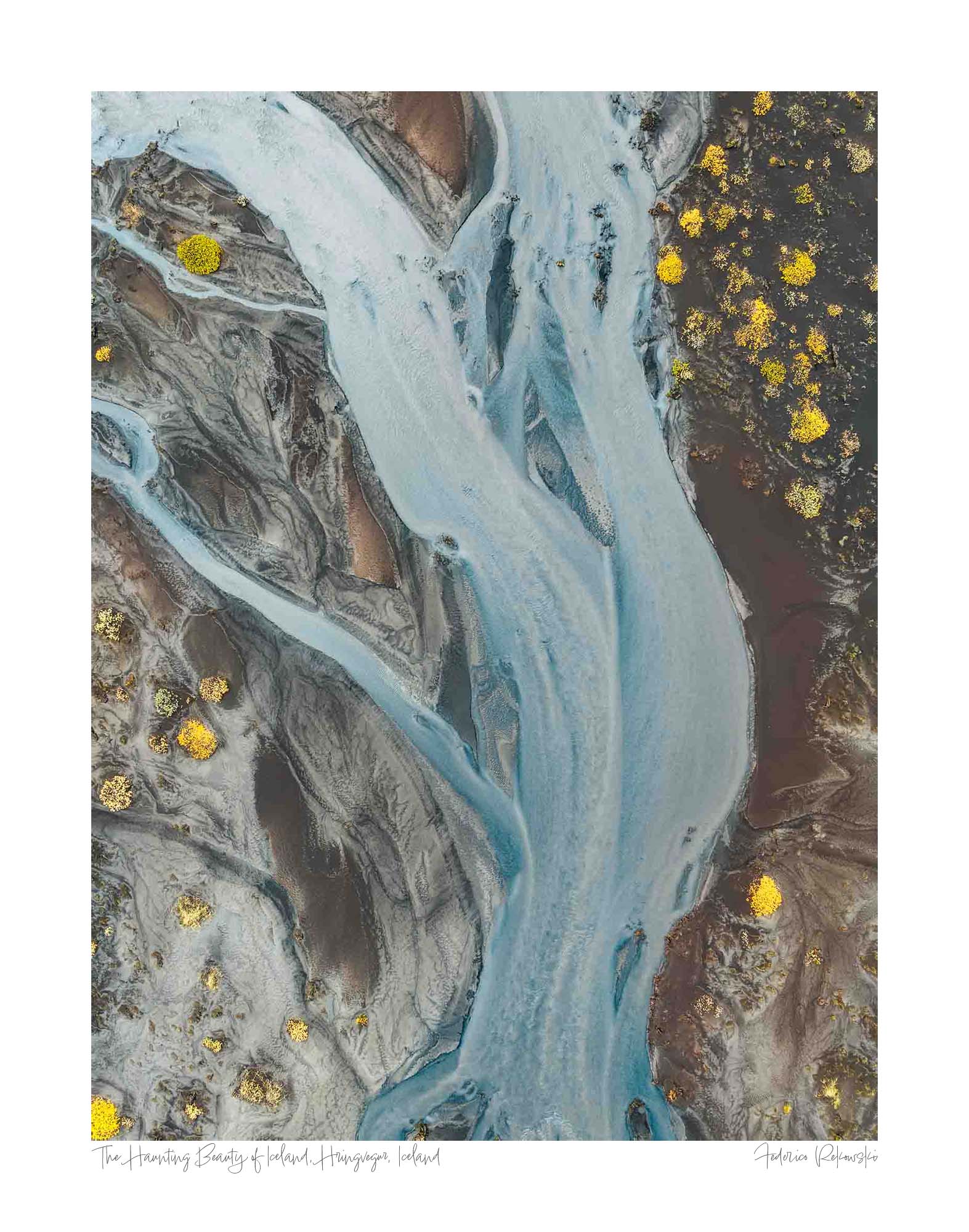 Aerial photo of a ghostly pale blue river snaking through the darker earth in Iceland, with patches of bright yellow flora.