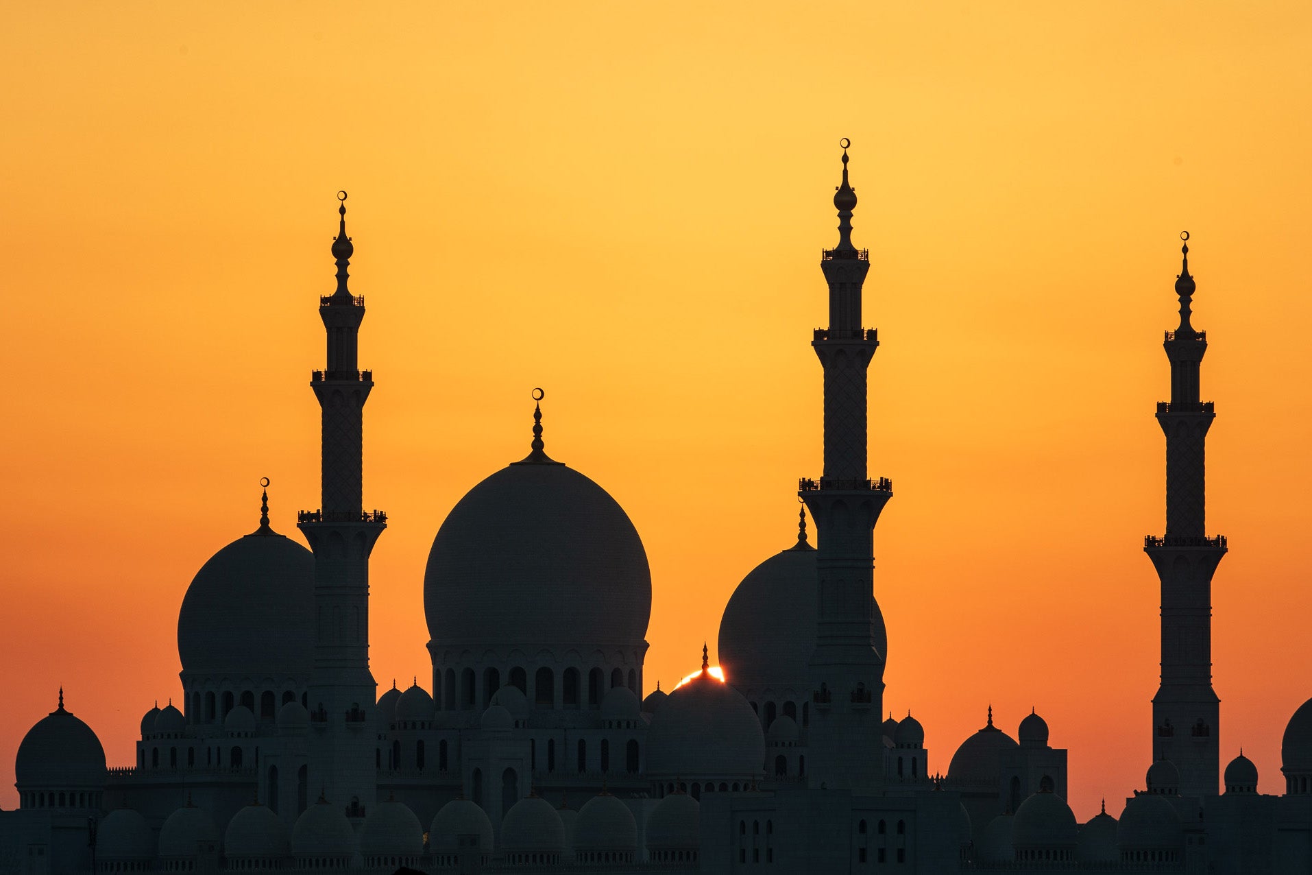 Silhouette of a grand mosque with multiple domes and minarets against a bright orange sunset sky in Abu Dhabi.
