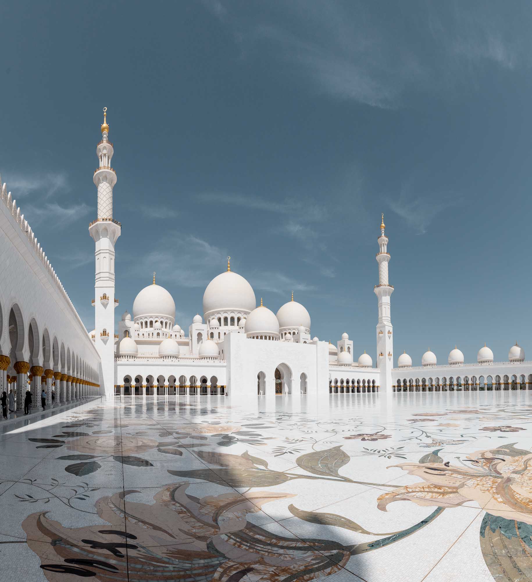 The Sheikh Zayed Grand Mosque in Abu Dhabi with its white marble domes and minarets, reflected on the glossy courtyard surface under a clear blue sky.