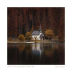 A small stone chapel with a spire, surrounded by trees in autumn colors and reflected in the calm waters of Lago di Braies, Dolomites.