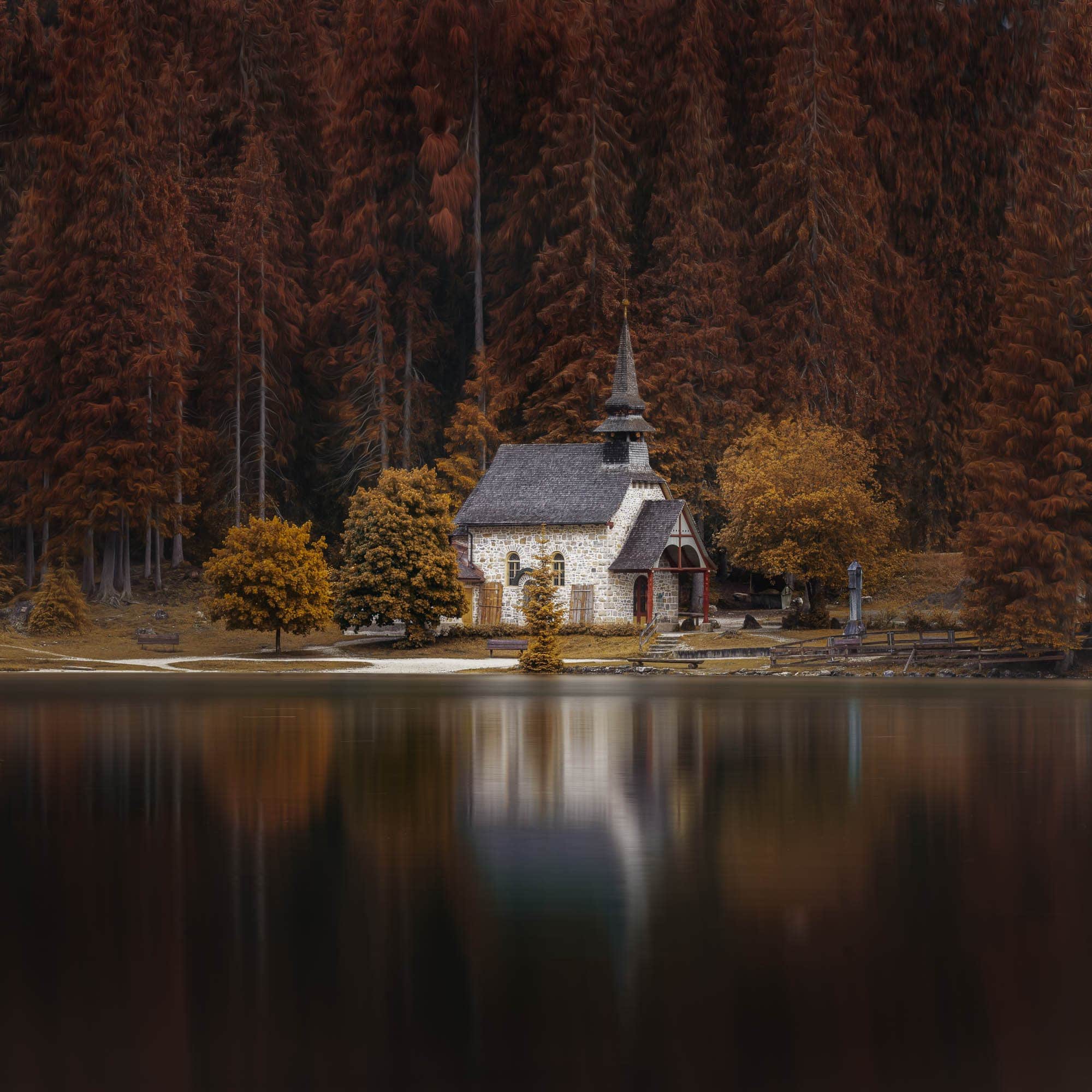 A small stone chapel with a spire, surrounded by trees in autumn colors and reflected in the calm waters of Lago di Braies, Dolomites.