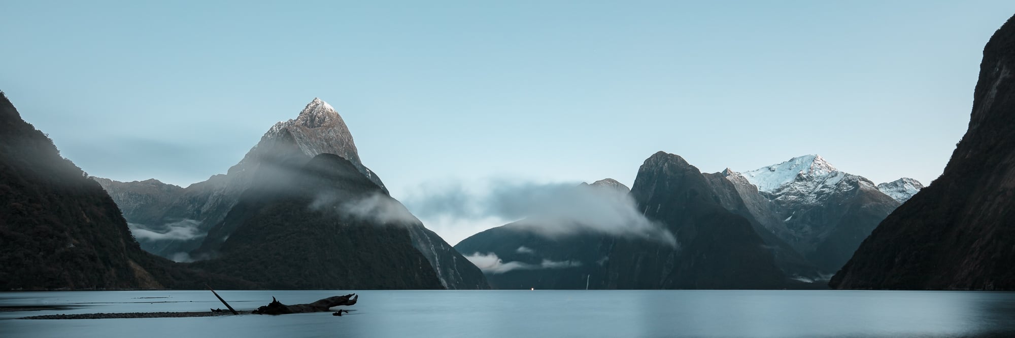 Panoramic view of Milford Sound in New Zealand, with the sunrise illuminating the mountain peaks and casting a tranquil light over the still waters.