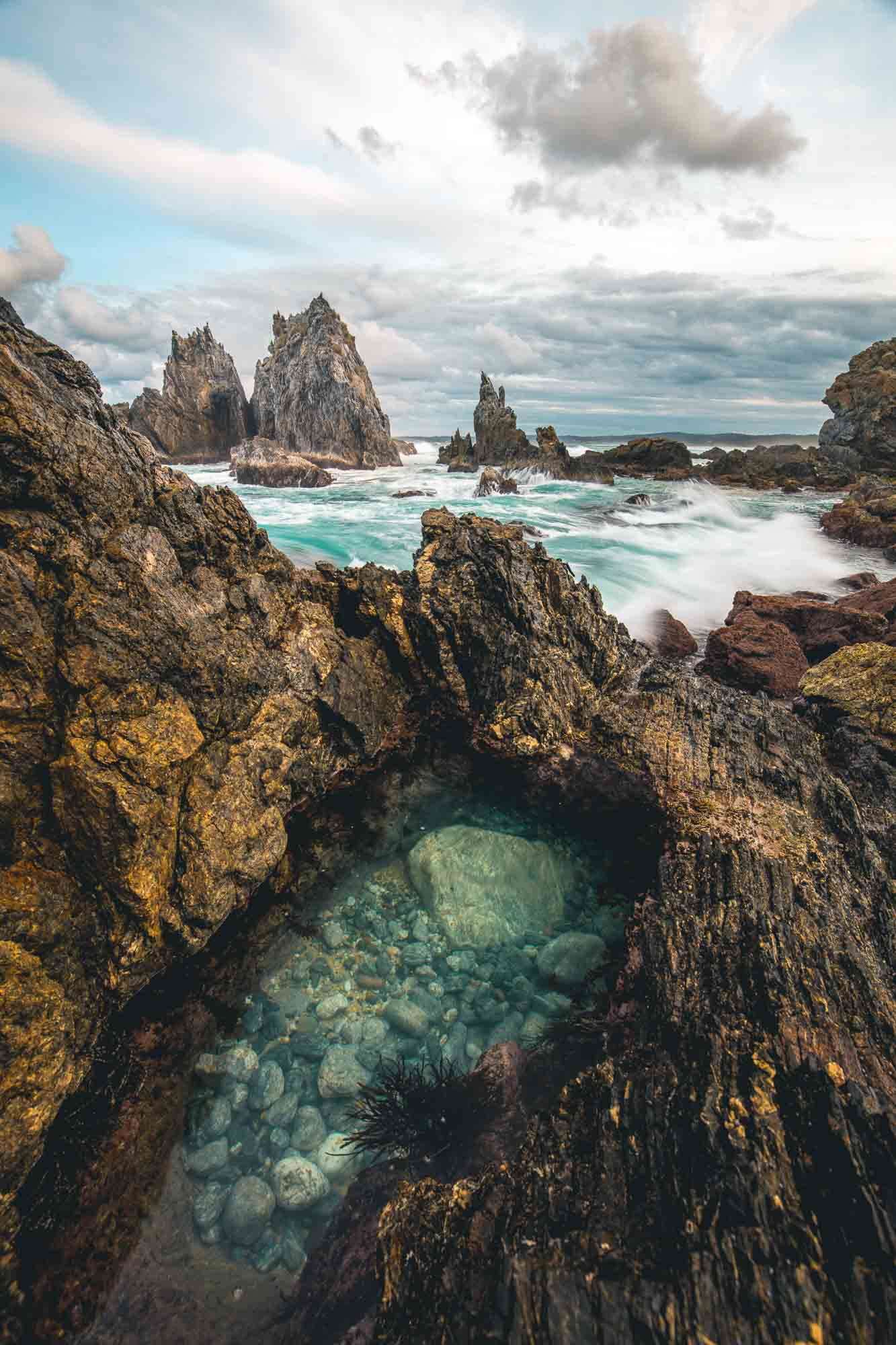 Rugged rock formations of Camel Rock in Bermagui with a natural tide pool in the foreground and turbulent sea in the background under a cloudy sky.
