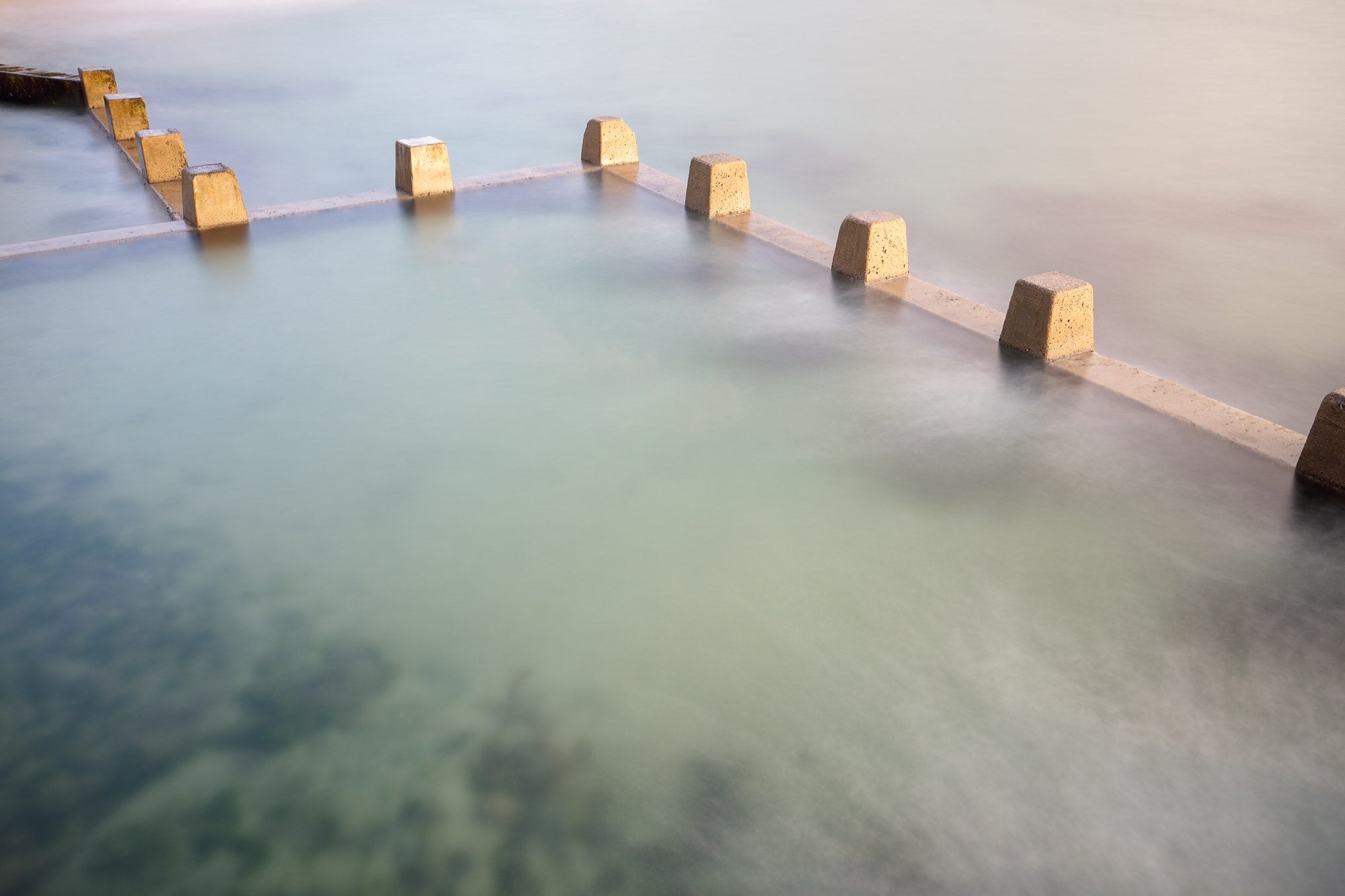 Long exposure photo of smooth, milky waters flowing around square concrete blocks in the ocean pool at Coogee Beach, Sydney, with subtle underwater details visible.
