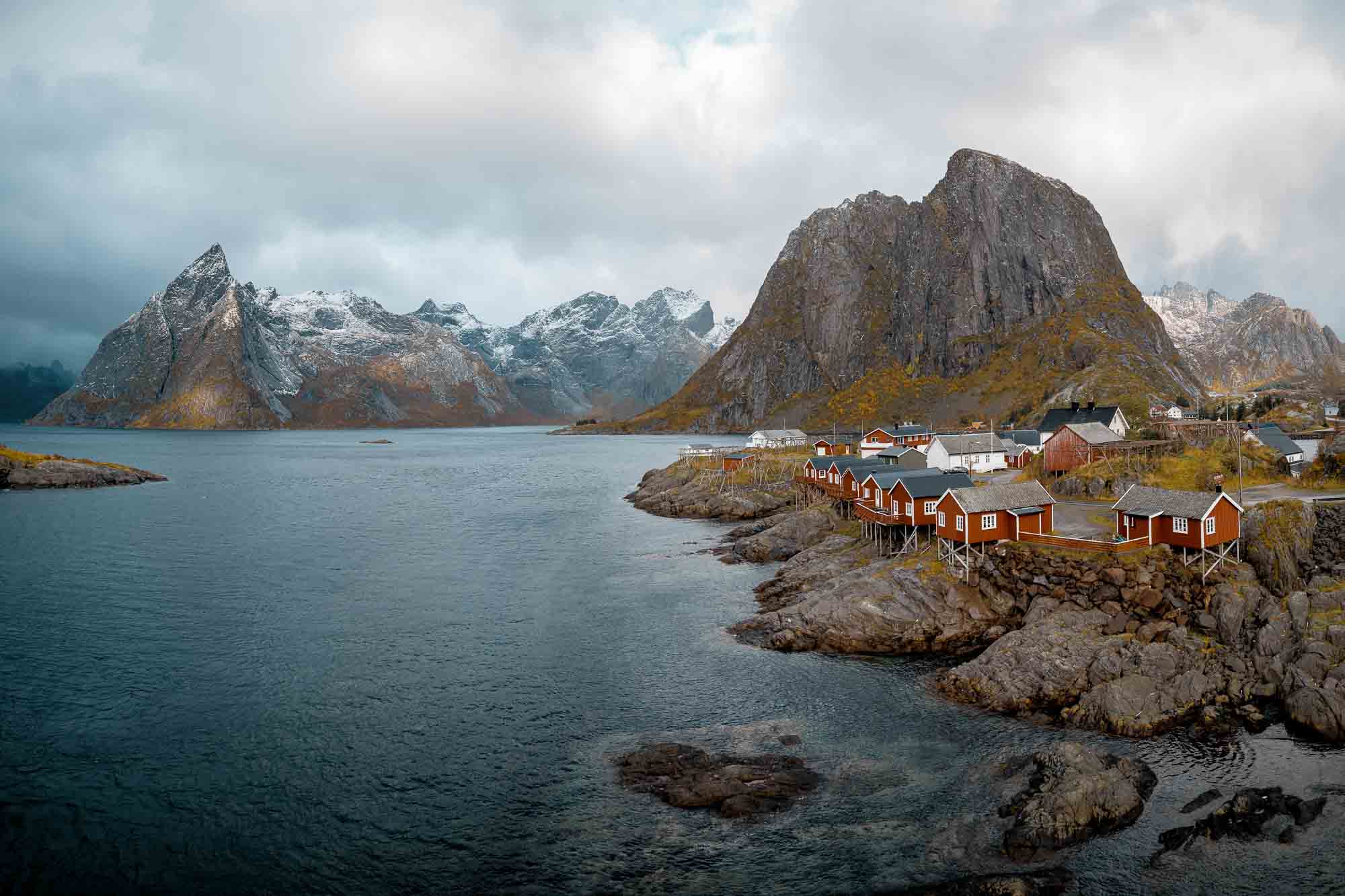 Red rorbuer cabins in Hamnøy, Lofoten Islands, Norway, with calm waters and snow-dusted mountains under an overcast sky.