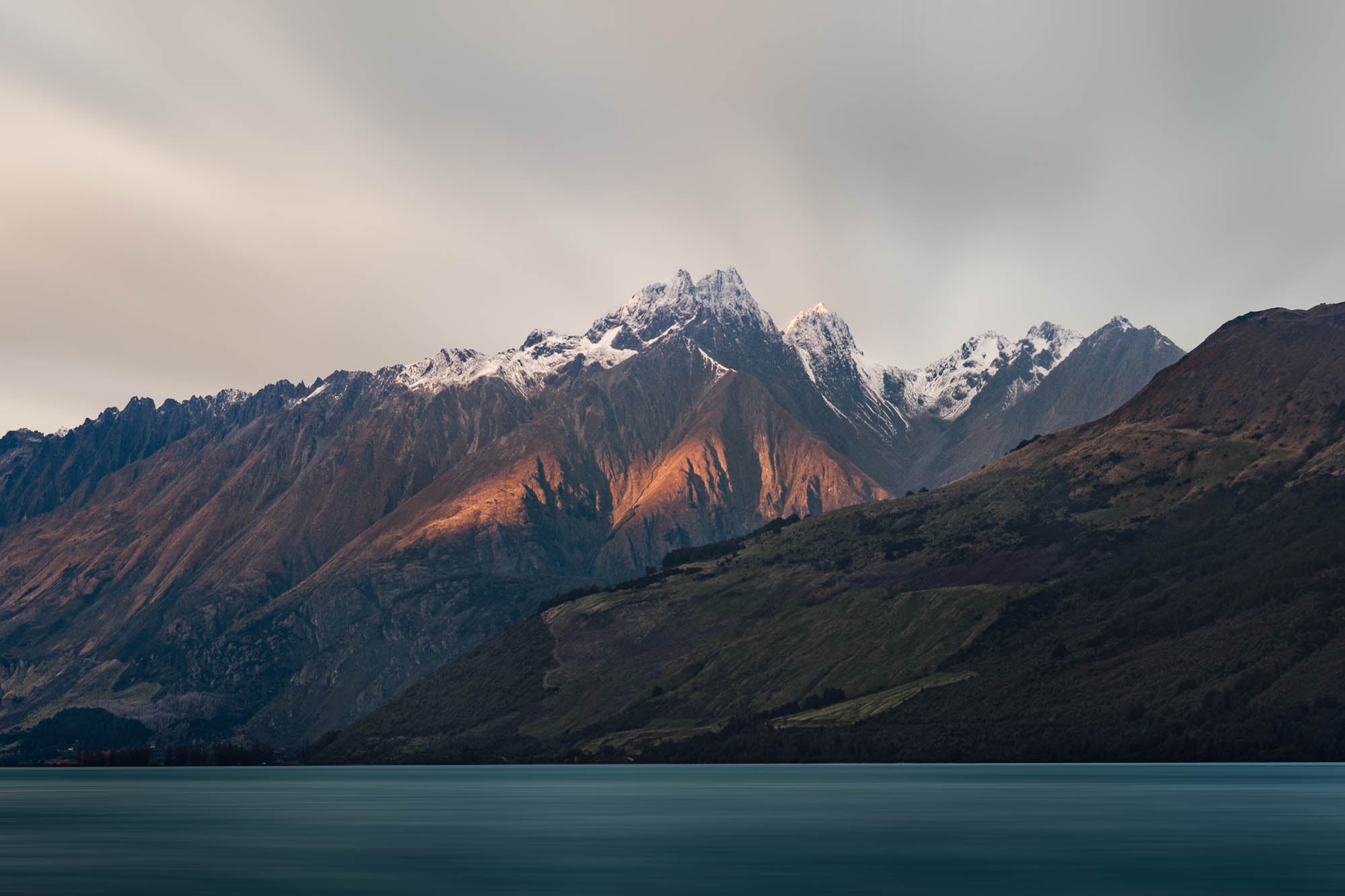 Last light of dusk casting a warm glow on the snowy peaks of mountains in Glenorchy, New Zealand, with a calm lake in the foreground.