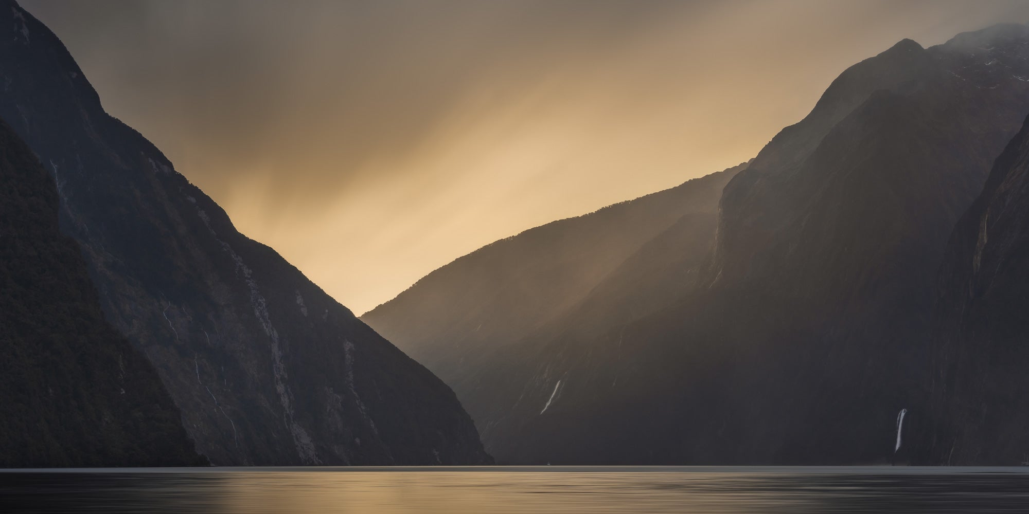 A sailboat sails on Milford Sound, New Zealand, with golden sunlight streaming through the mountainous valley.