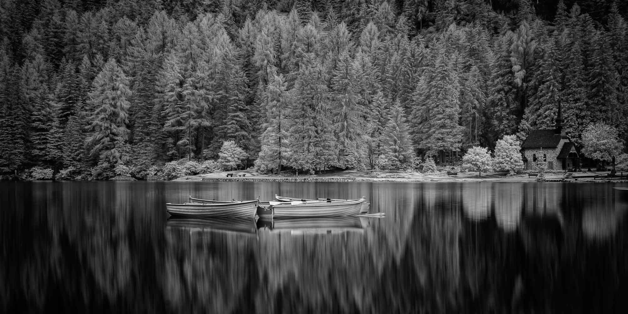 Black and white image of Lago di Braies with still water, reflective boats, a small chapel, and dense trees in the background.