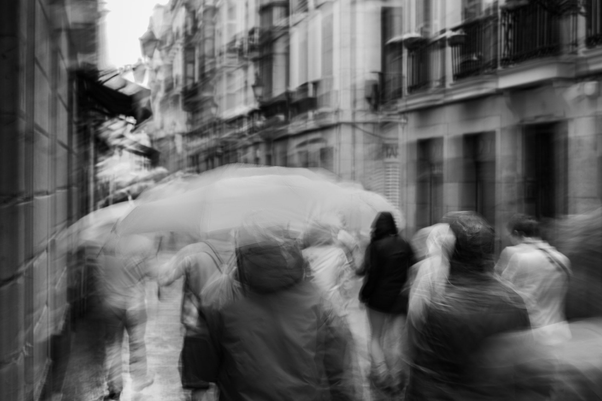 A monochrome image of pedestrians with umbrellas blurred by motion, embodying rain's lullaby in San Sebastian, inspired by Langston Hughes’ poetry.