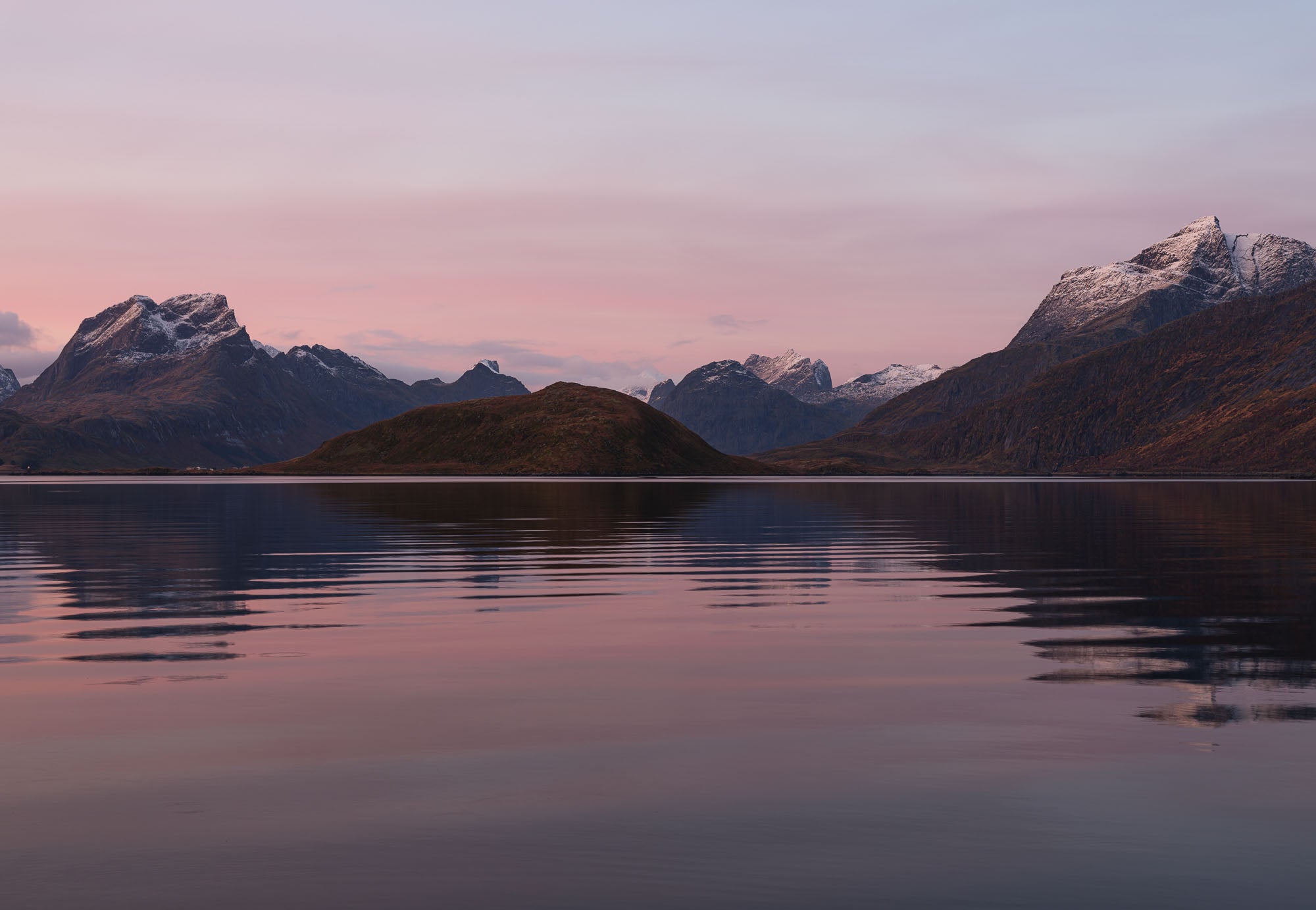 Sunrise at Fredvang in Lofoten, with snow-capped mountains reflecting in calm waters under a pastel-hued sky.