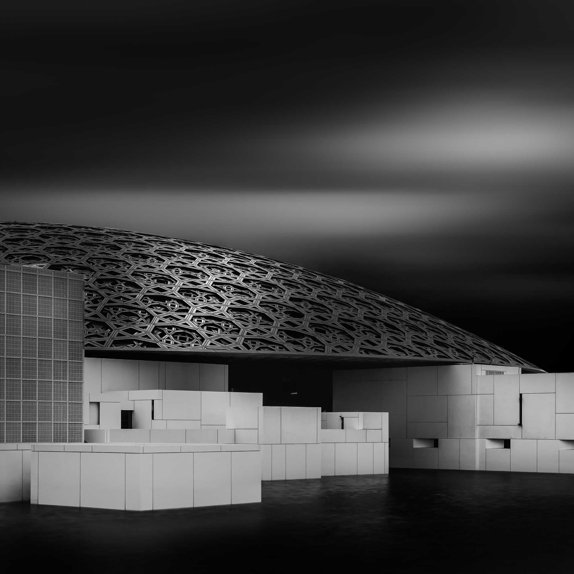 Black and white photo of the Louvre Abu Dhabi, highlighting the intricate geometric patterns of the dome against a dramatic sky.