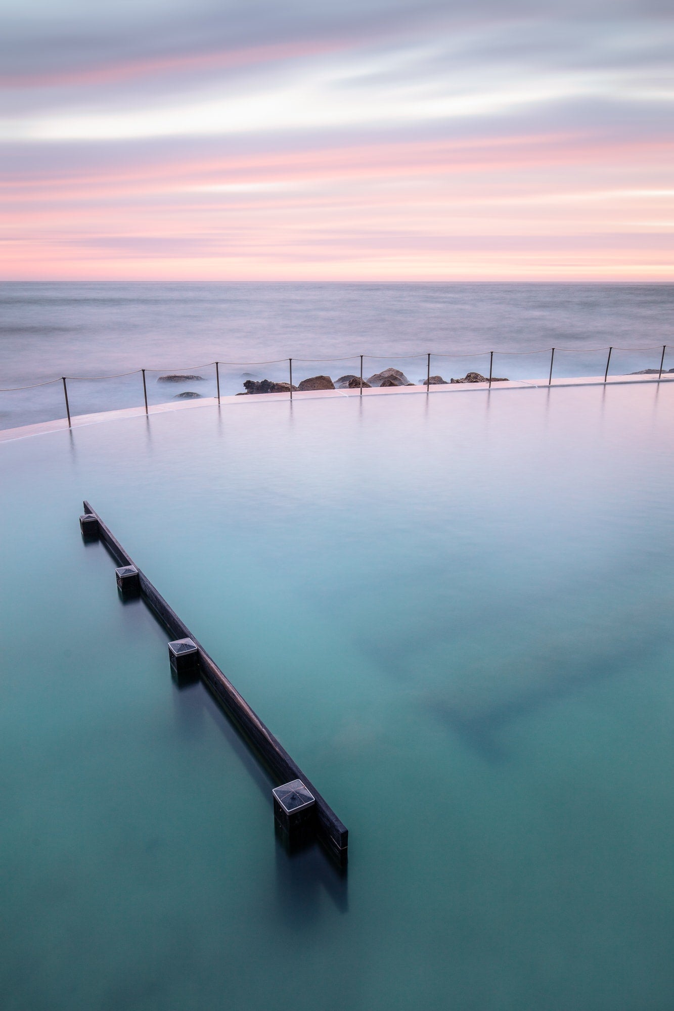 Long exposure of Bronte Baths in Sydney, with smooth waters under a pink and blue striped sky at twilight.