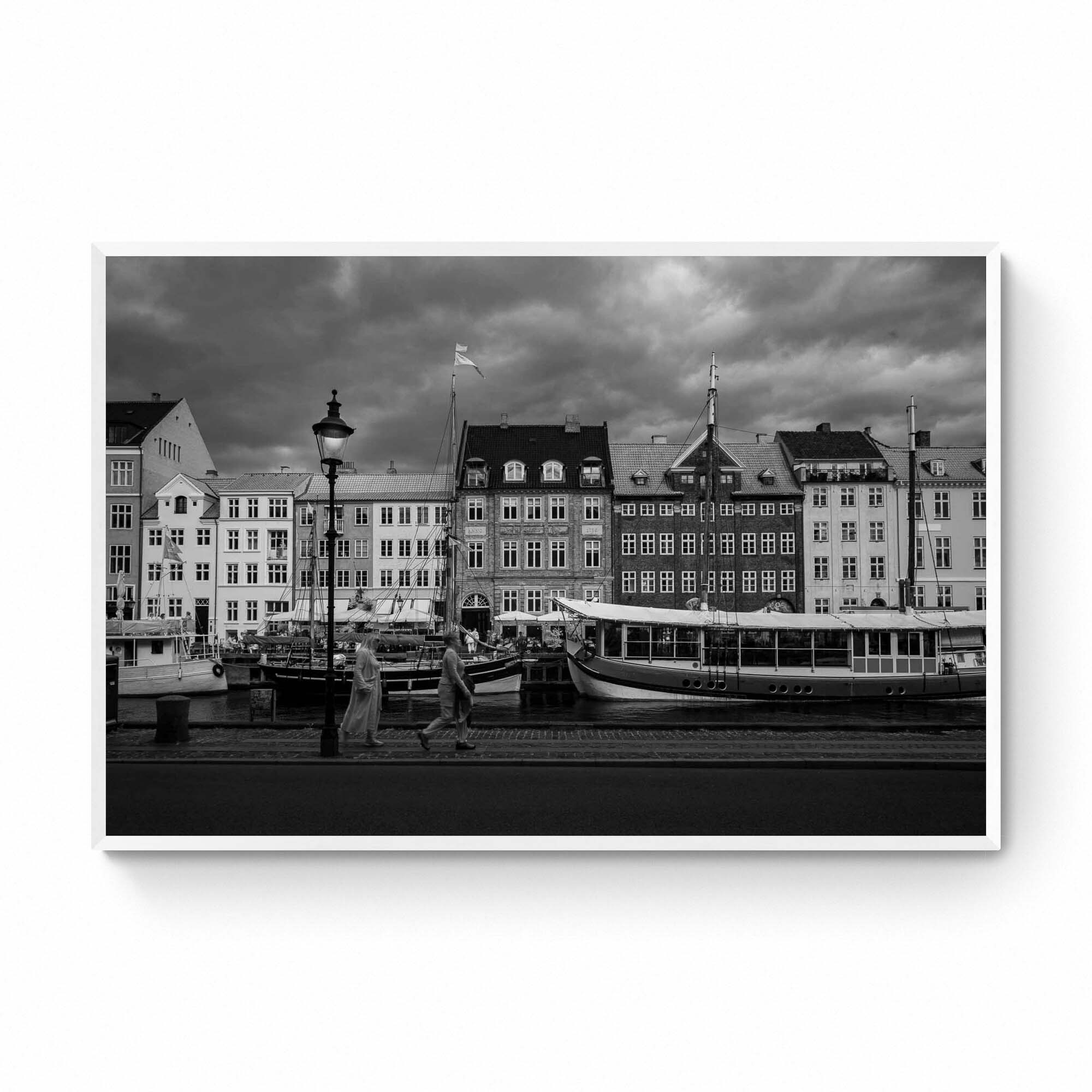 Black and white image of Nyhavn, Copenhagen, with historical buildings along the waterfront, boats docked in the canal, and people walking by.