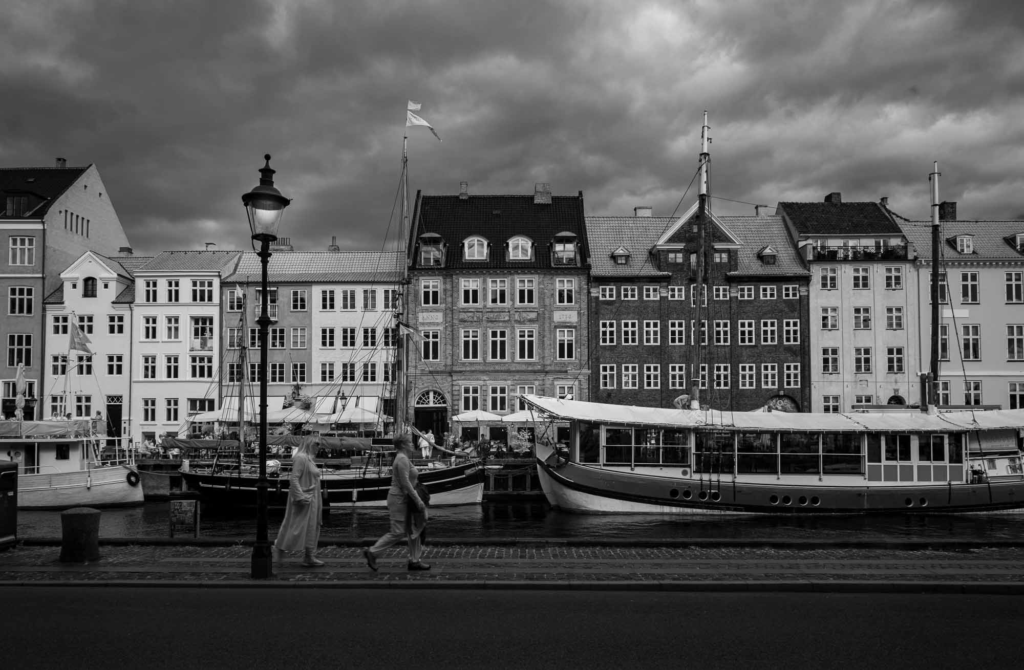 Black and white image of Nyhavn, Copenhagen, with historical buildings along the waterfront, boats docked in the canal, and people walking by.