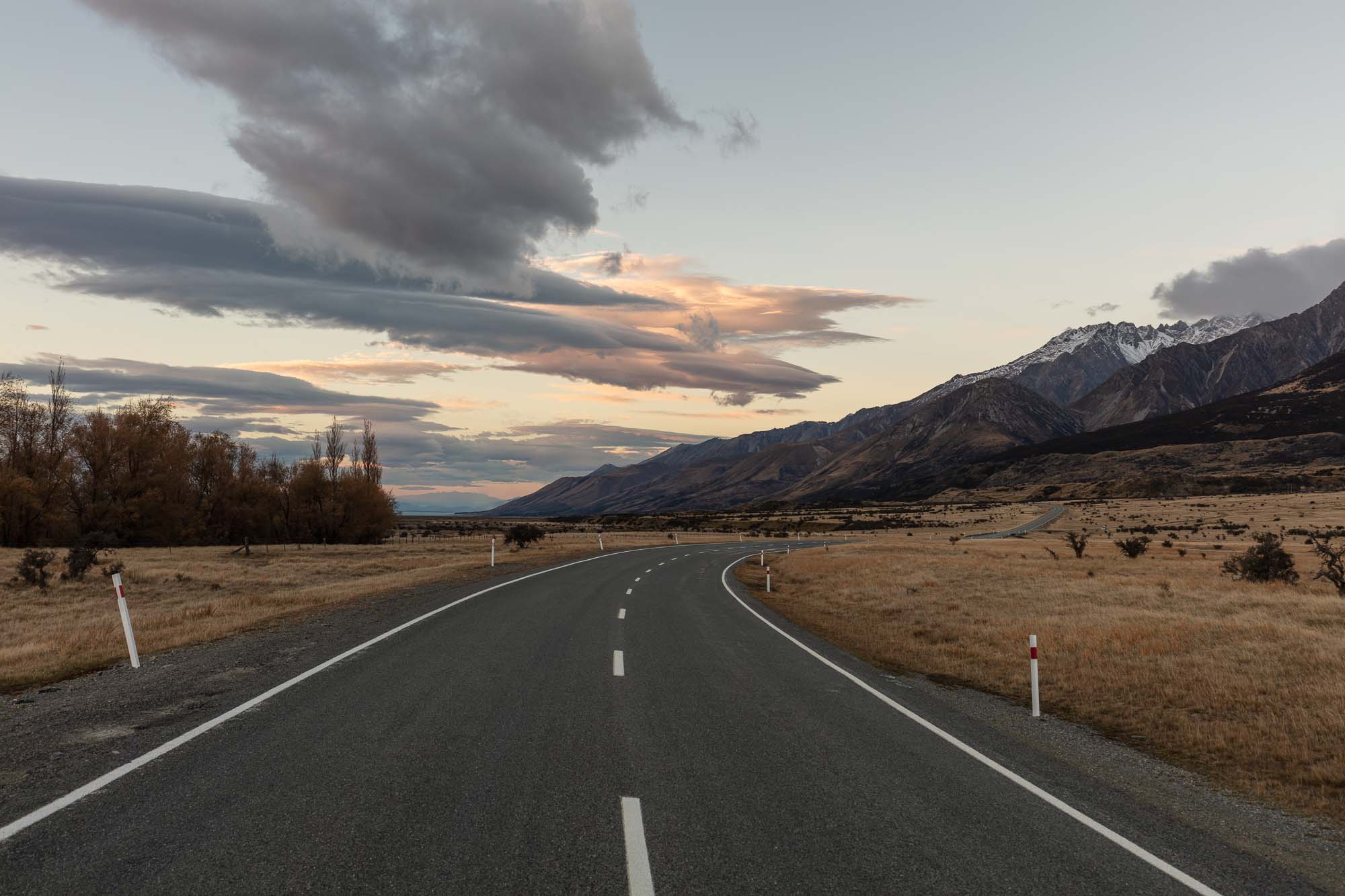 A serene landscape photograph of a road in New Zealand with Aoraki/Mt Cook in the background under a twilight sky with dynamic cloud formations.