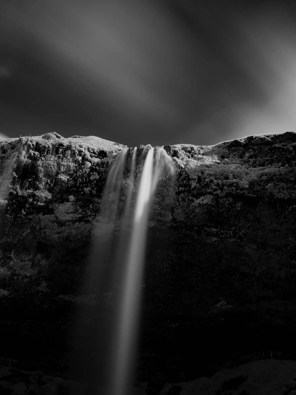 A black and white long-exposure photograph of Seljalandsfoss waterfall in Iceland, showing smooth water streams against a dark, textured cliff.