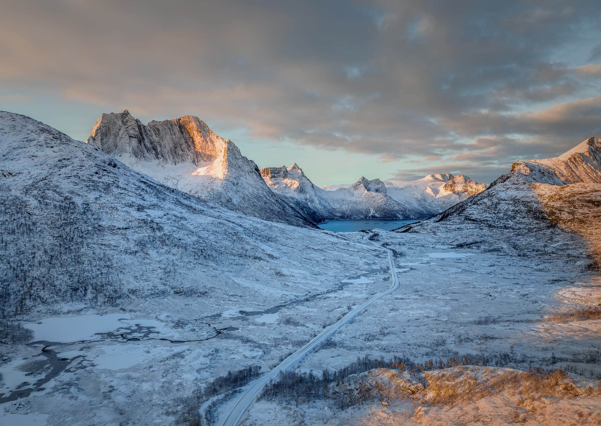 Snow-covered landscape at Husøy, Senja, with mountains glowing in the soft light of the setting sun and a road winding through the valley.