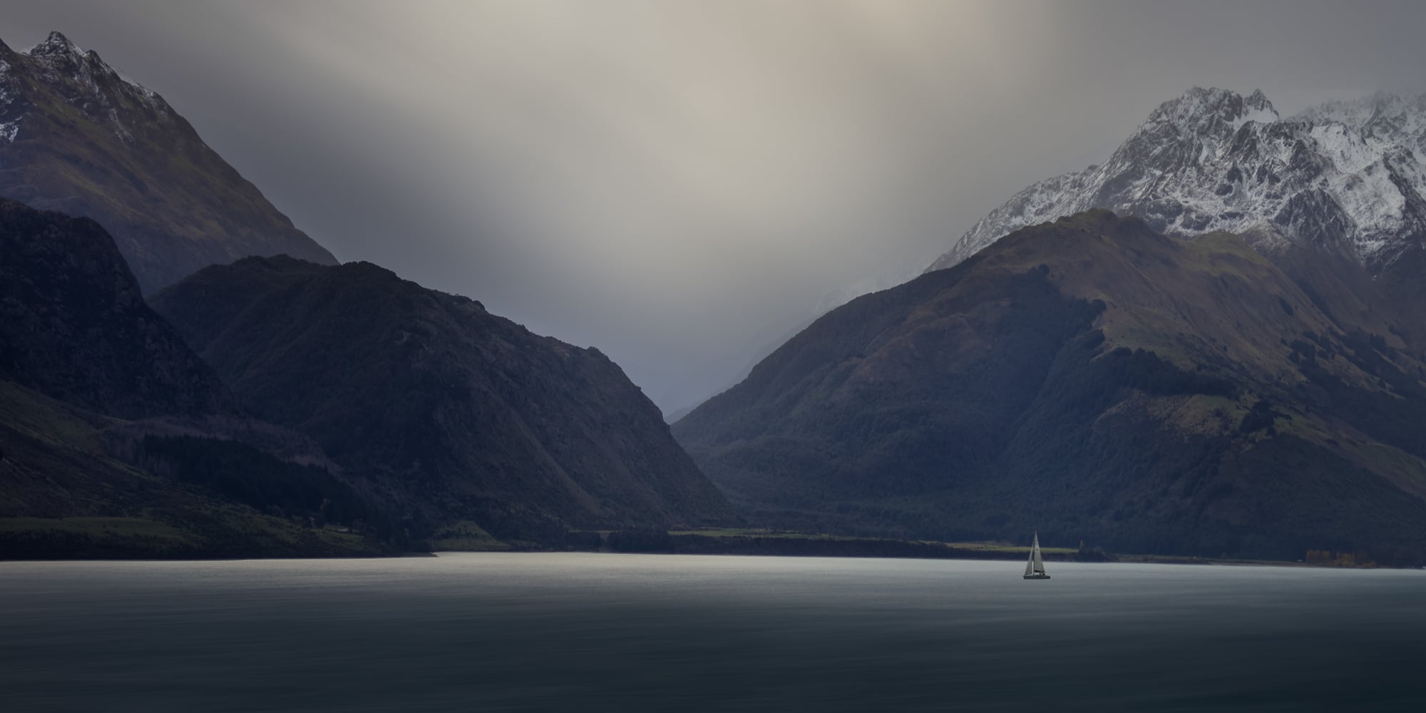 A lone sailboat on a calm lake in Queenstown, New Zealand, with towering, snow-capped mountains under a soft, overcast sky.
