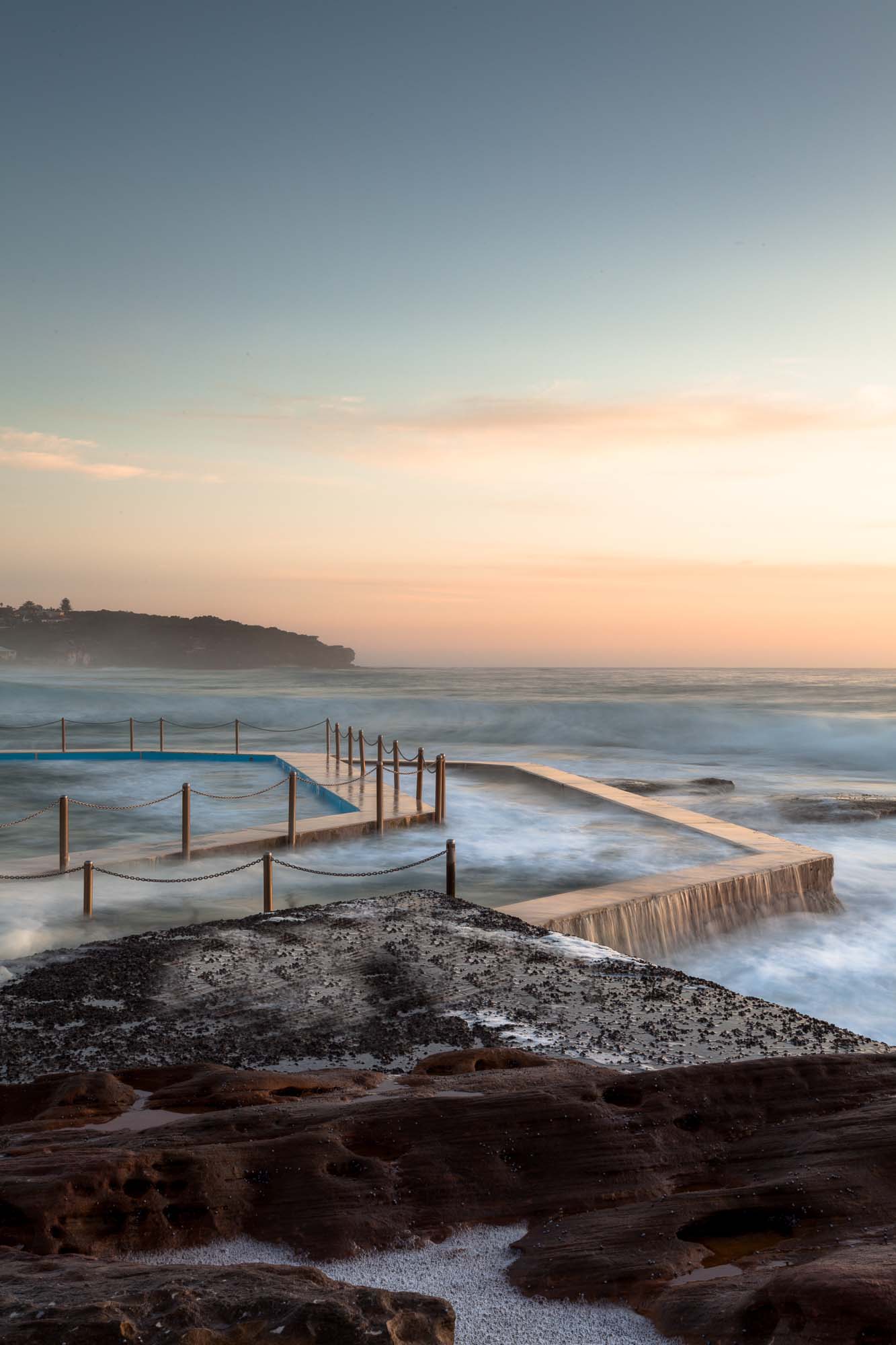 Twilight at South Curl Curl pool with smooth water flowing over its edges, bordered by textured rocks against a pastel sky.