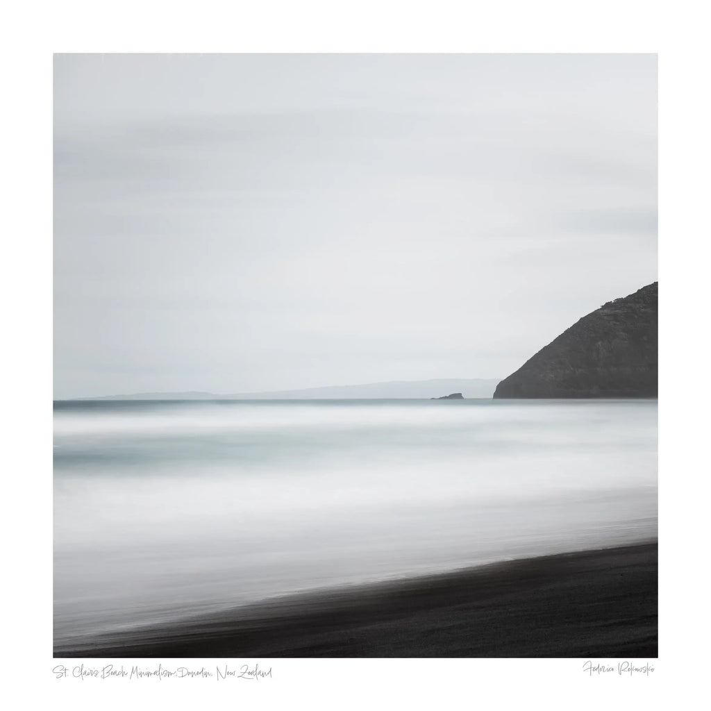 A minimalist long exposure photo of the calm sea meeting the dark sands of St. Clair's Beach in New Zealand, with a headland silhouette on the right.