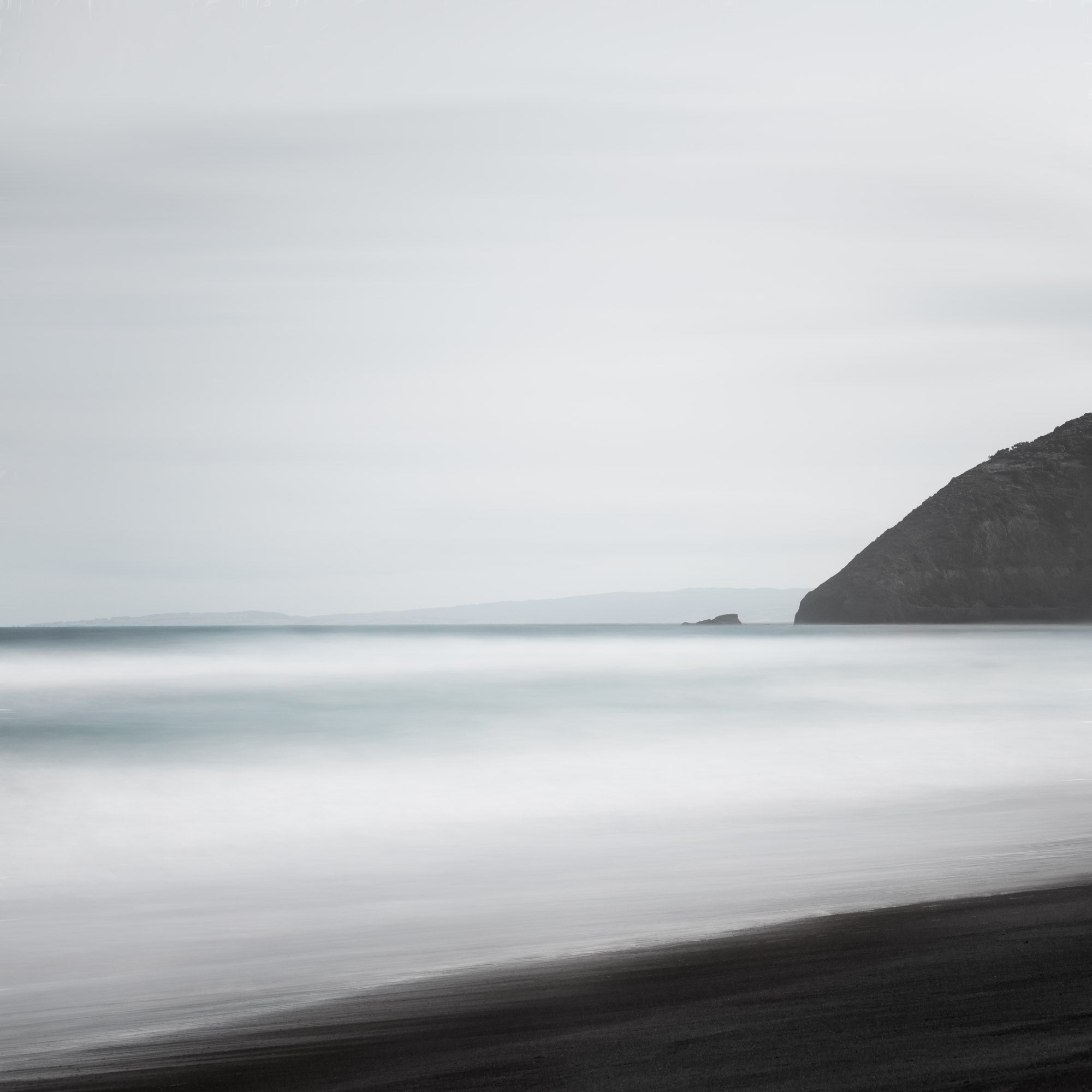 A minimalist long exposure photo of the calm sea meeting the dark sands of St. Clair's Beach in New Zealand, with a headland silhouette on the right.