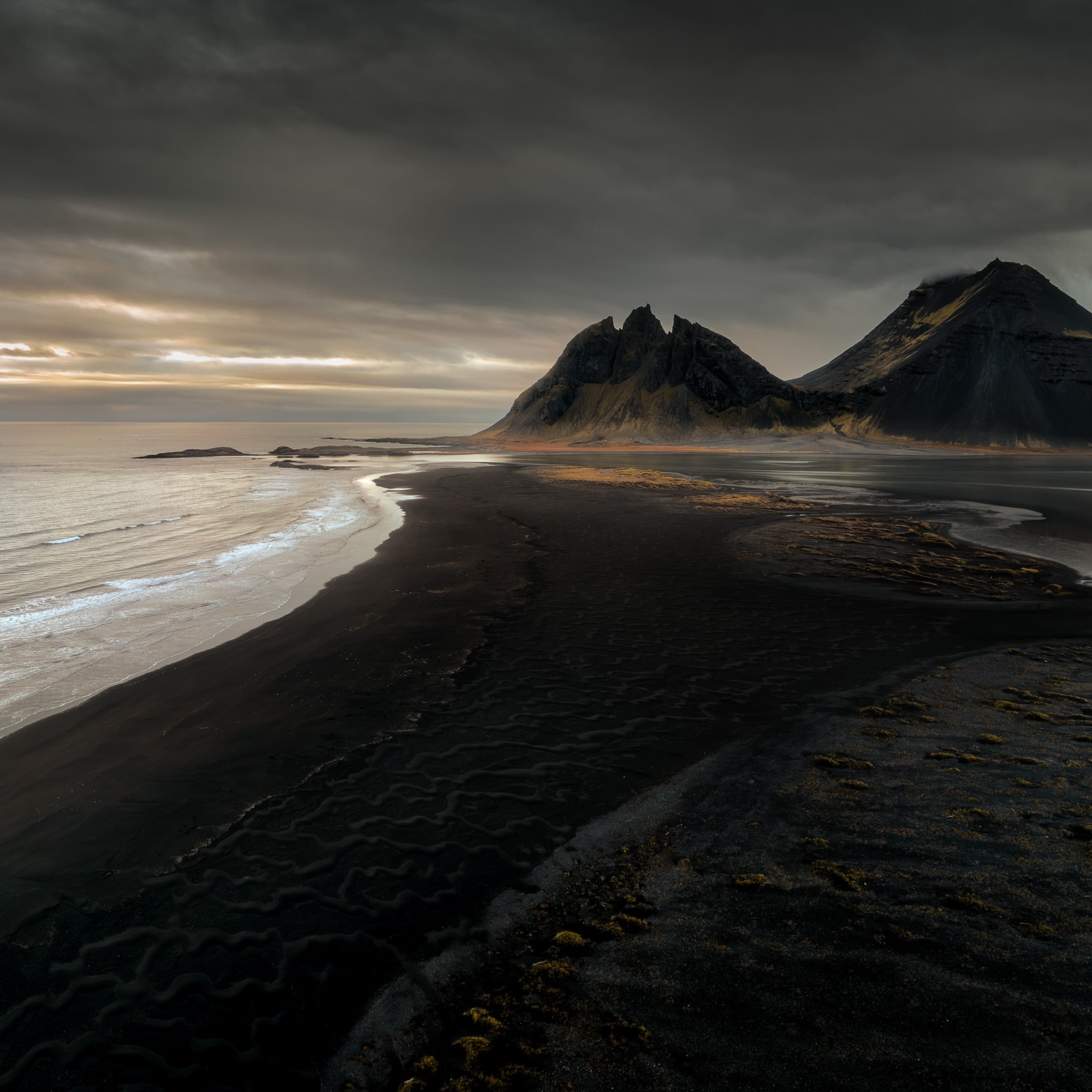Sunset at Vestrahorn, Iceland, with the mountain's silhouette against a dramatic sky and patterns in the dark sand leading to the sea.