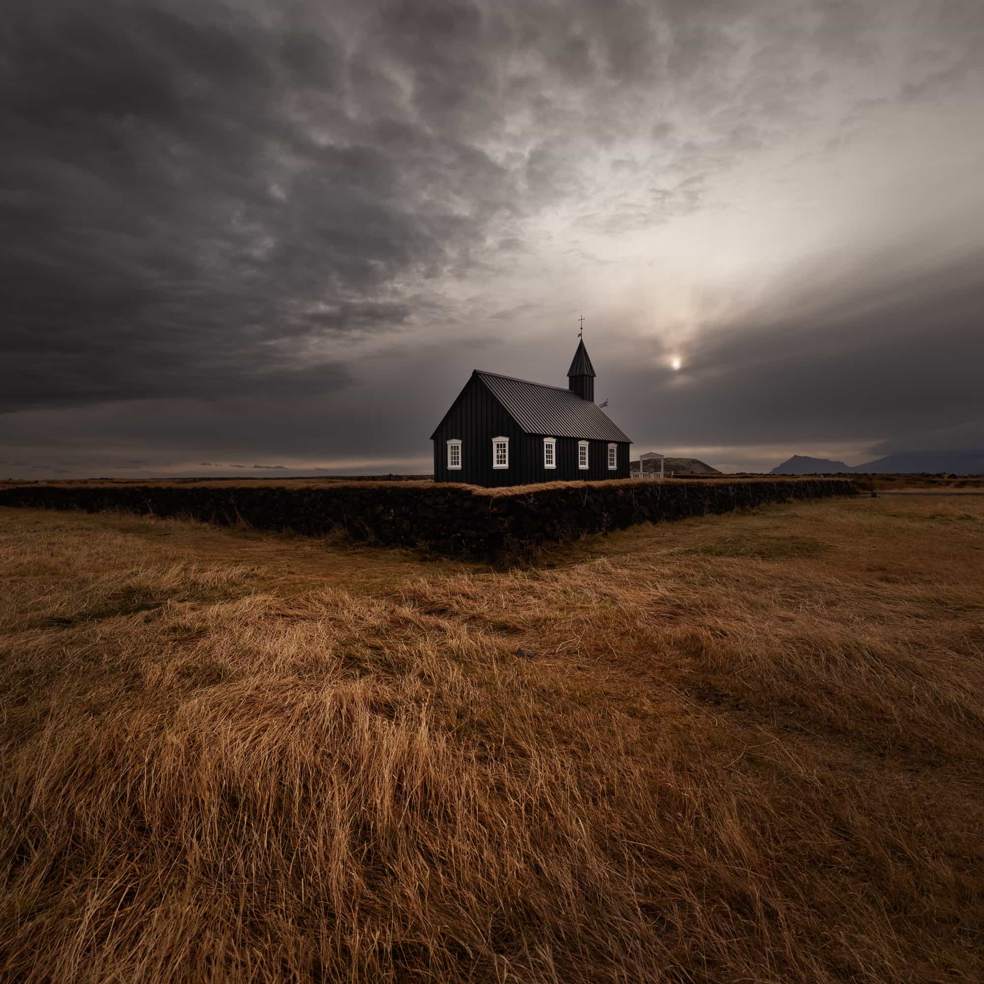 A dark, solitary church in the open Icelandic landscape with dry grass in the foreground and a dramatic, cloud-filled sky above, capturing a moment of quietude before a storm.