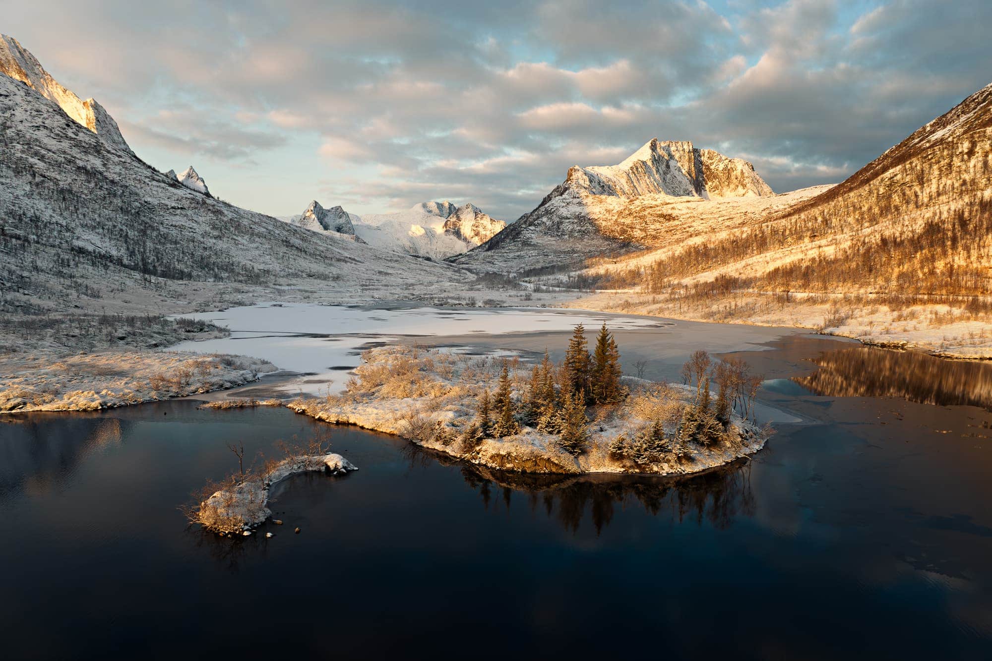 Sunset illuminating the snowy peaks and frozen lake of Holvatnet in Senja, Norway, with frosted trees on an islet.