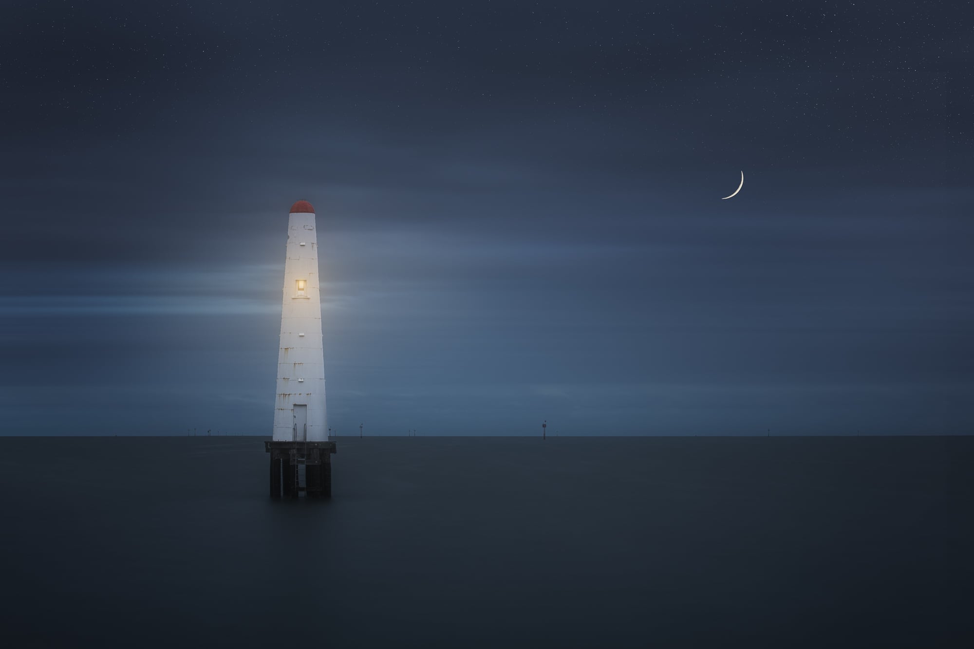A lone lighthouse illuminated on Princess Pier in Melbourne against a starry night sky with a crescent moon.