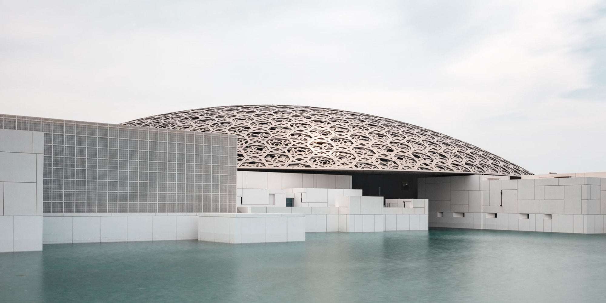 Wide-angle view of the Louvre Abu Dhabi’s intricate dome over calm water, under a soft sky.
