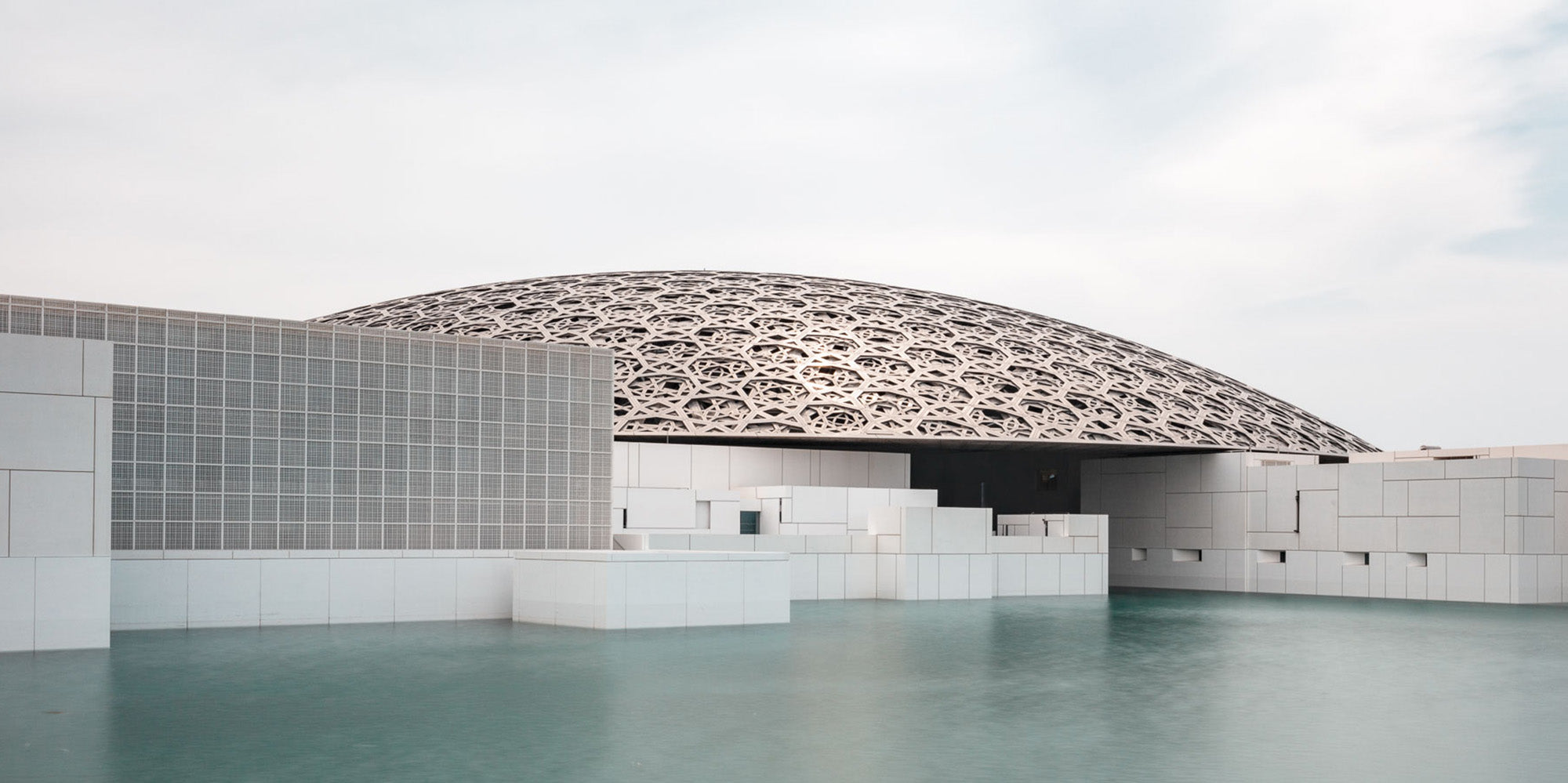 Wide-angle view of the Louvre Abu Dhabi’s intricate dome over calm water, under a soft sky.