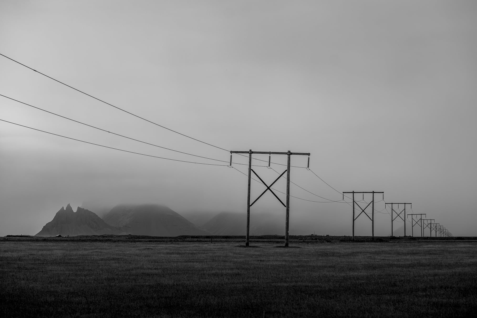Black and white photo of power lines running across a field in Hofn, Iceland, with mountains faintly visible in the misty background.