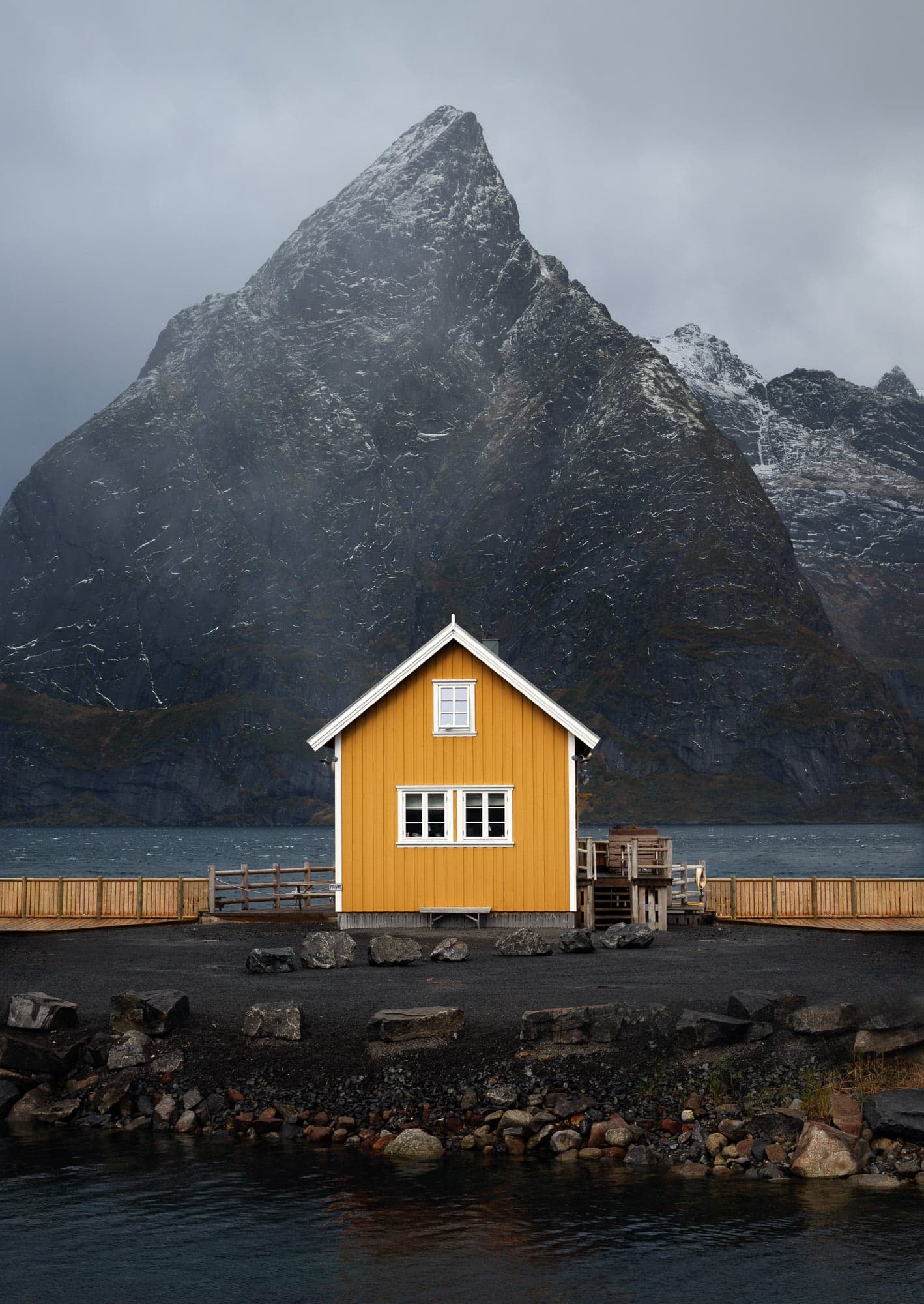 A solitary yellow cabin stands in the foreground with a towering, snow-streaked mountain rising dramatically behind it in Lofoten, Norway.