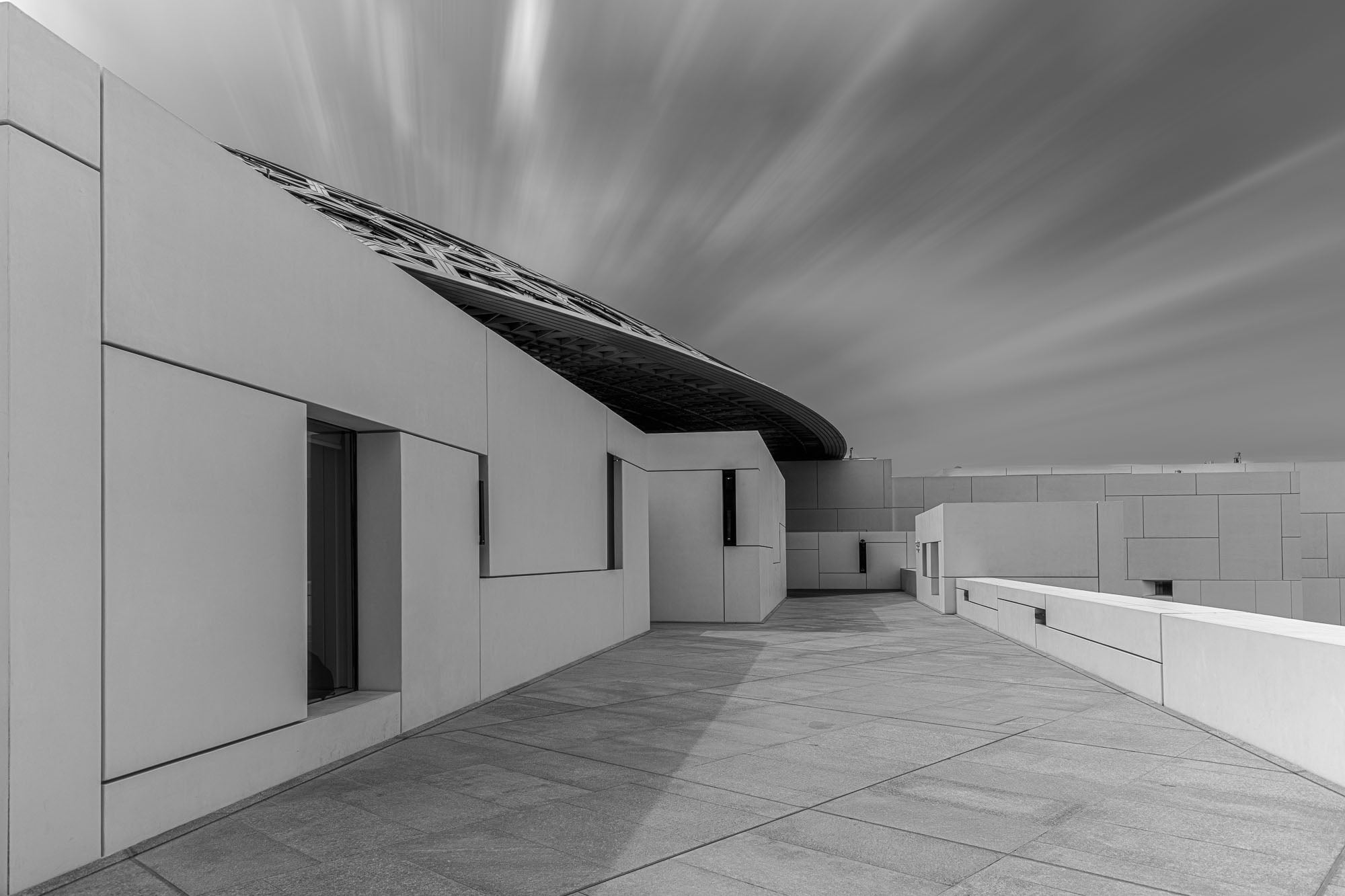 A long exposure black and white image of the Louvre Abu Dhabi showing the sharp angles of the museum's architecture against the smooth streaks of clouds in the sky.