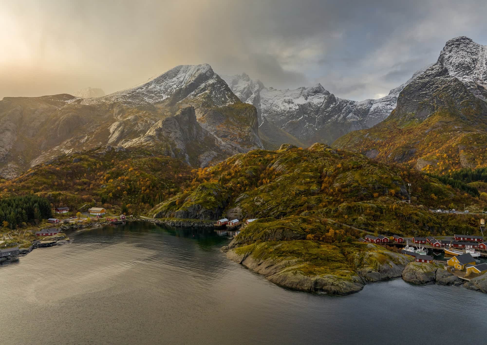 Aerial view of a fjord in Lofoten, Norway, during autumn with snow-capped mountains and colorful houses nestled amongst golden-hued trees.
